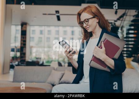 Young happy businesswoman in glasses with laptop and agenda under her arm, looking at smartphone, checking last e-mails or financial company reports w Stock Photo