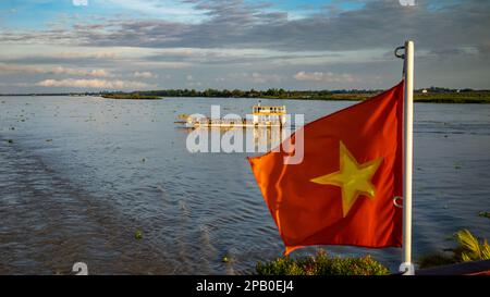 A crowded river ferryboat crosses the Mekong River past a Vietnamese flag on the stern of the river cruise ship Victoria Mekong just after dawn downst Stock Photo