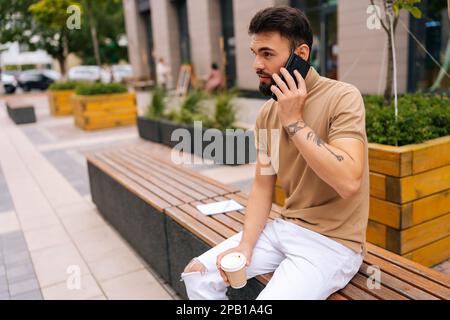 Handsome tattooed young man talking on smartphone holding in hand takeaway coffee sitting on bench on city street. Bearded male having conversation Stock Photo