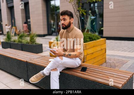 Thoughtful male artist with tattooed hands sitting on bench with takeaway coffee and making sketches with pen on notebook, looking away. Stock Photo
