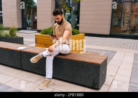 Wide shot of focused tattooed man sitting on bench and using typing smartphone looking to screen on city street. Stock Photo