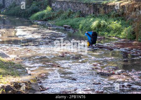 Zapopan, Jalisco Mexico. January 1, 2023. Stream with a homeless person sitting on stones washing his clothes, flowing water, wild green plants and st Stock Photo