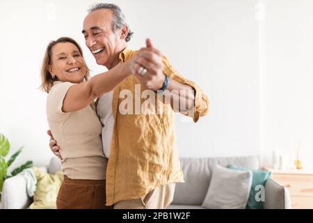 Cheerful retired spouses husband and wife dancing and laughing in living room, happy romantic couple enjoying slow dance Stock Photo