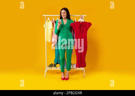 App for online shopping. Young european lady using cellphone and holding dress, ordering clothes online in mobile app Stock Photo