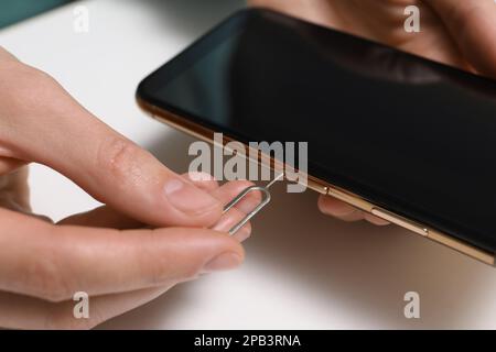 Woman with ejector opening SIM card tray in smartphone at white table, closeup Stock Photo