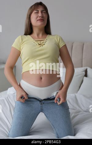 Young woman struggling to squeeze into tight jeans on bed at home Stock Photo