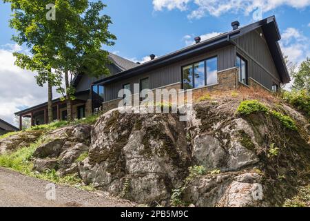 Cut stone and charcoal wood plank with black trim hybrid timber frame home built on top of outcrop in summer. Stock Photo