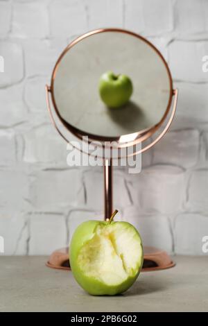 Bitten green apple near mirror with reflection of whole fruit on table Stock Photo