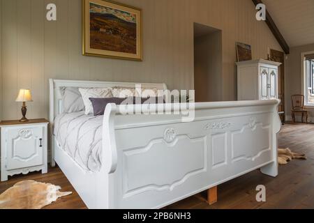 White painted king size sleigh bed in main bedroom with brushed wood plank walls and red pine wood floorboards inside hybrid timber frame home. Stock Photo
