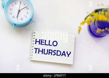 Hello Thursday words on a notepad and a bouquet of mimosa flowers in a blue vase on a white table with a clock. Happy Thursday concept. Template for a Stock Photo