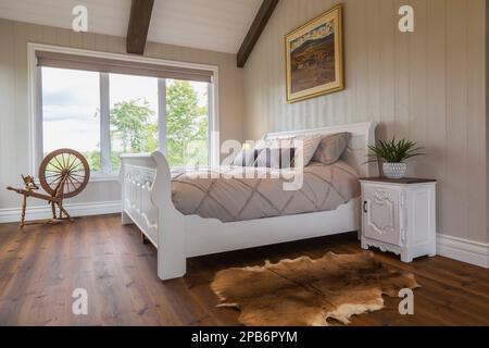 White painted king size sleigh bed in main bedroom with brushed wood plank walls and red pine wood floorboards inside hybrid timber frame home. Stock Photo
