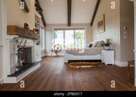 White painted king size sleigh bed in main bedroom with decorative fireplace, brushed wood plank walls, red pine wood floorboards inside timber home. Stock Photo