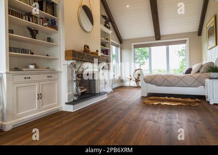 White painted king size sleigh bed in main bedroom with decorative fireplace, brushed wood plank walls, red pine wood floorboards inside timber home. Stock Photo