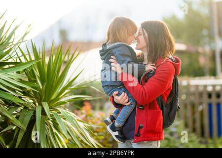 Cute funny toddler boy in his mothers arms. Mom and son having fun on sunny spring day in a park. Adorable son being held by his mommy. Stock Photo