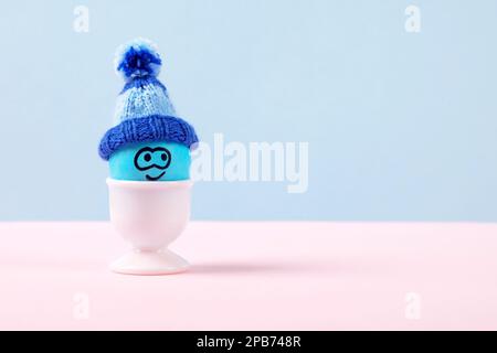 One blue Easter egg with funny face and crocheted hat in white stands on a pink table. Happy Easter concept. Greeting card. Stock Photo