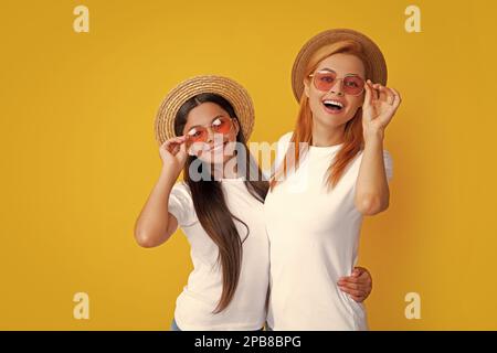 Happy stylish mother and daughter posing at studio yellow background, wearing straw hat and sunglasses. Summer family portrait. Stock Photo