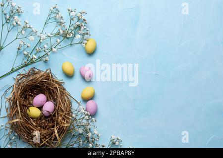 Banner for the Easter holiday. Colored eggs in a straw nest and white spring flowers on a blue background. Place for text. Top view. Stock Photo