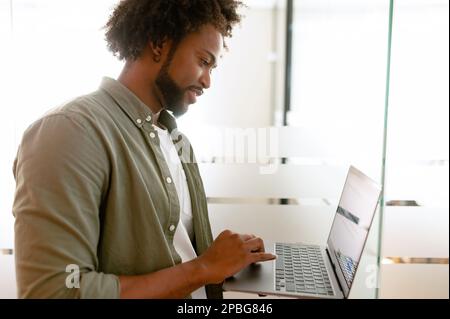 Optimistic ethnic man in casual wear using laptop sitting in modern open space office, young african-american male employee replying emails, working on a new project, side view Stock Photo