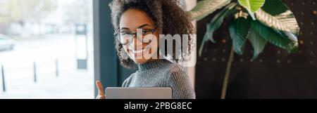 Smiling woman holding laptop and looking at camera while standing near window in cafe Stock Photo