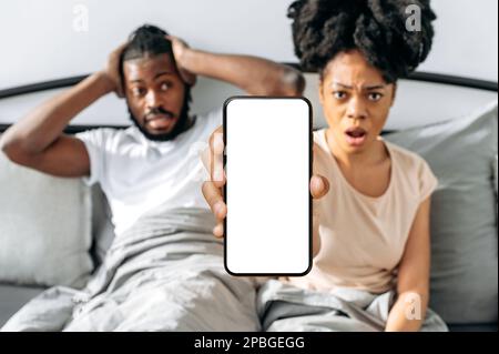 Shocked puzzled african american couple, sit in a cozy bed in the bedroom, woman shows smartphone with blank mock-up screen, space for advertising, presentation, look at camera at a loss Stock Photo