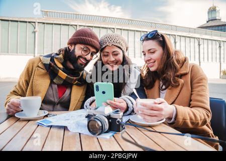 Group of young friends having fun watching videos on a social media app using a cellphone device it a coffee shop terrace. Three happy people smiling taking a selfie with a smart phone outdoors. High quality photo Stock Photo