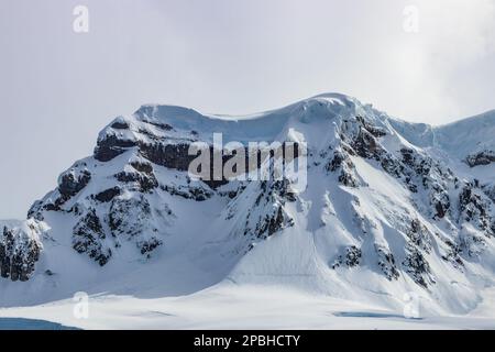 Rugged mountain peak on Antarctic Peninsula. Steep slopes covered with snow; underlying rocks exposed. Blue ice visible. Cloudy sky in background. Stock Photo