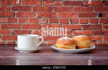 A cup of hot black coffee and a plate with two fresh donuts in powdered sugar on a wooden table against a red brick wall. Close-up. Stock Photo