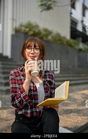 Portrait, Charming young Asian female college student in casual clothes sipping coffee while reading book on the stairs outside of the campus building Stock Photo