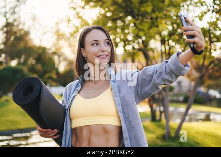 Shot of sporty woman holding a yoga mat taking a selfie in the park after a workout, smiling and leading a healthy lifestyle. The concept of a healthy Stock Photo