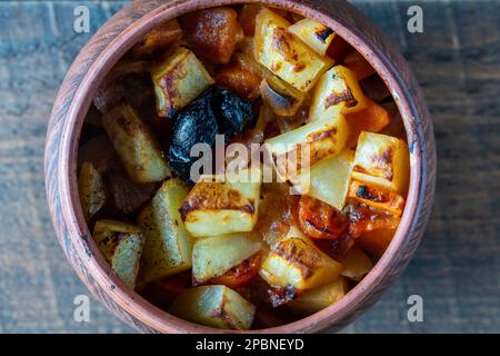 Stewed potato, carrot, onion, tomato and prunes in a clay pot on wooden table background, close up, top view Stock Photo