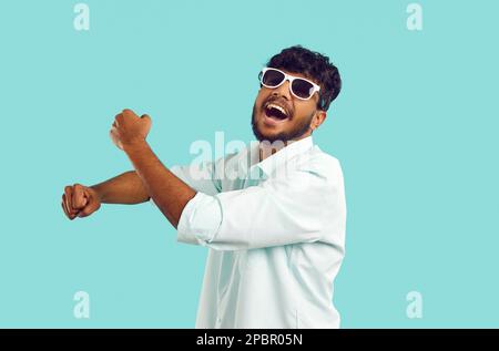 Cheerful young Indian man laughing, singing, having fun and dancing against light blue background. Stock Photo