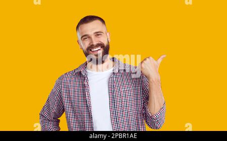 Cheerful bearded handsome Caucasian man pointing with thumb to side at advertising space. Stock Photo