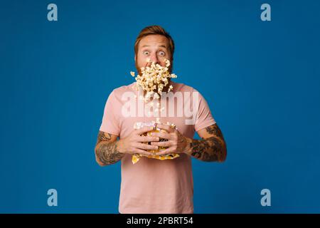 Shocked bearded white man in casual wear holding bucket with flying popcorn isolated over blue studio background Stock Photo