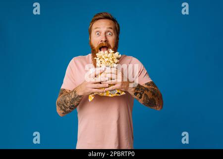 Shocked bearded white man in casual wear holding bucket with flying popcorn isolated over blue studio background Stock Photo