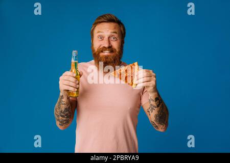 Bearded happy man holding pizza slice and beer while standing isolated over blue background Stock Photo