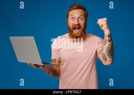 Bearded happy man screaming and holding laptop while standing isolated over blue background Stock Photo