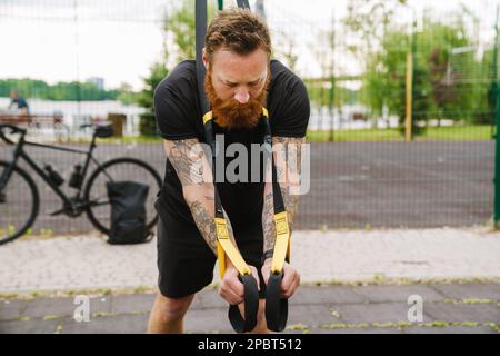 Young handsome tattooed bearded serious man doing exercises with TRX near bicycle at outdoor sports field Stock Photo