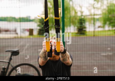 Cropped portrait of young tattooed bearded man doing exercises with TRX near bicycle at outdoor sports field Stock Photo