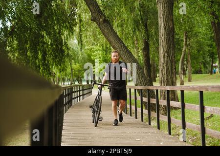 Adult handsome tattooed bearded man in black sport suit looking aside, while walking with bicycle in park Stock Photo