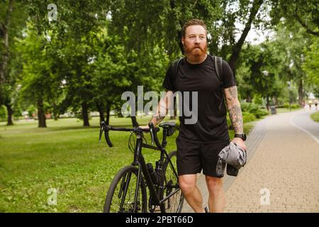 Adult handsome tattooed bearded man in black sport suit looking aside, while walking with bicycle in park Stock Photo