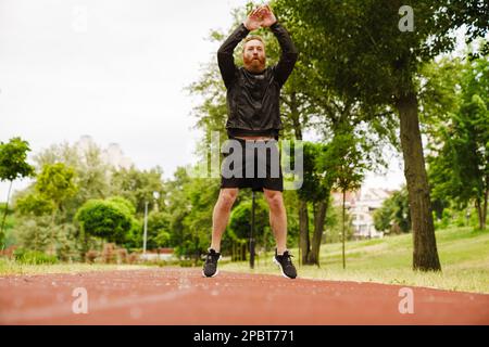Adult handsome bearded redhead man jumping in the air with raised arms and looking aside, while standing on running track outdoors Stock Photo