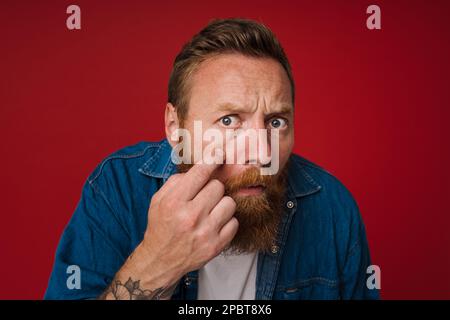 Adult bearded tattooed handsome stylish redhead man touching his cheek and looking at camera like in a mirror, while standing over isolated red backgr Stock Photo