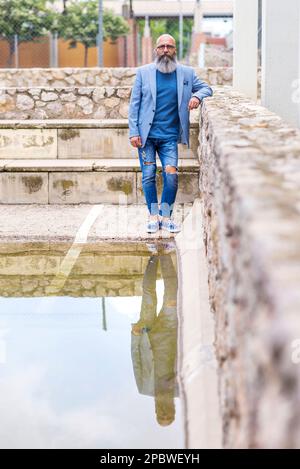 bald, bearded man standing leaning against a park wall by the water. Stock Photo