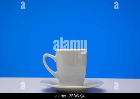 Small white ceramic coffee cup on a plate. Close up studio shot, isolated on blue background. Stock Photo