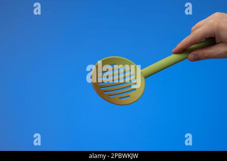 Green plastic heat-resistant perforated strainer ladle held by Caucasian male hand. Close up studio shot, isolated on blue background. Stock Photo