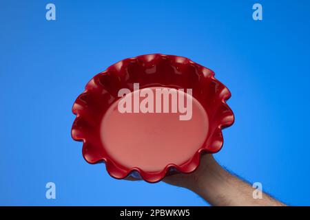 Round empty red ceramic pie tray  held by Caucasian male hand. Close up studio shot, isolated on blue background. Stock Photo