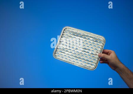 Empty single use metal aluminum foil cooking tray held by Caucasian male hand. Close up studio shot, isolated on blue background. Stock Photo