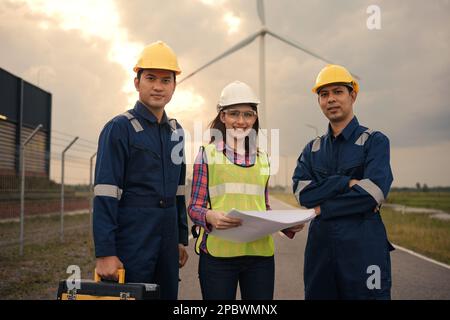 Happy three technician engineer in uniform with standing and checking wind turbine power farm power generator station. Stock Photo