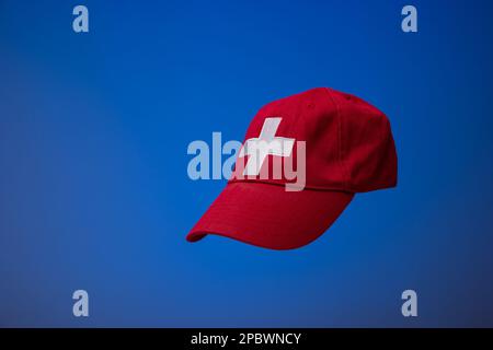 Red baseball cap with Swiss flag white cross. Close up studio shot, isolated on blue background. Stock Photo