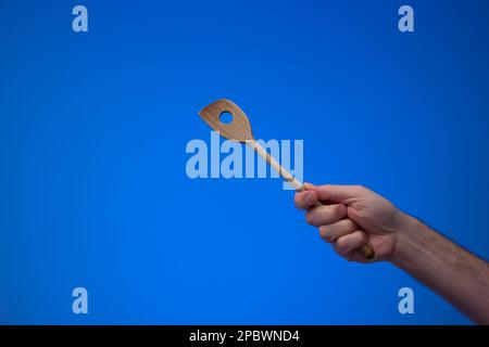 Used wooden kitchen spoon with long handle held in hand by man. Close up studio shot, isolated on blue background. Stock Photo